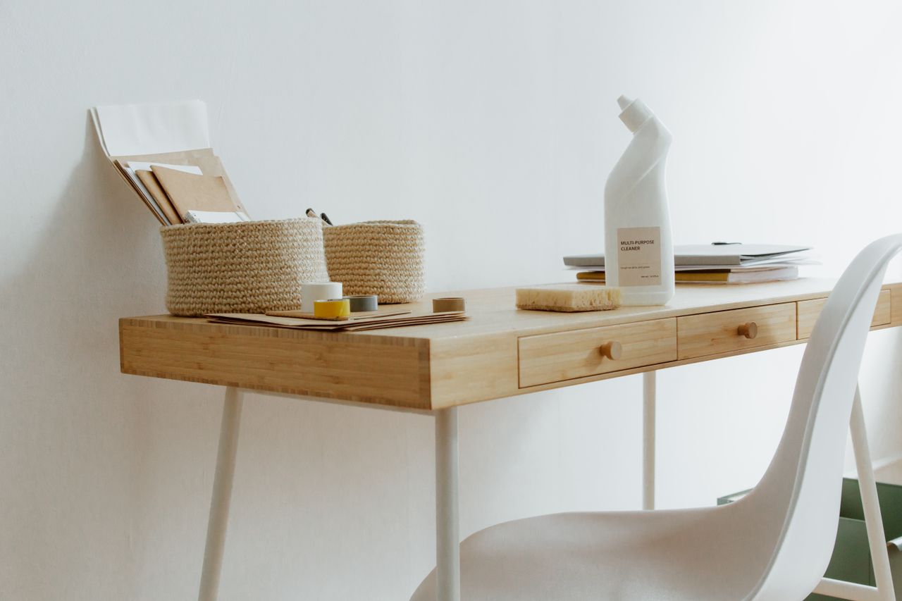A tidy, well-arranged home office desk featuring cleaning supplies, paperwork, and storage baskets.